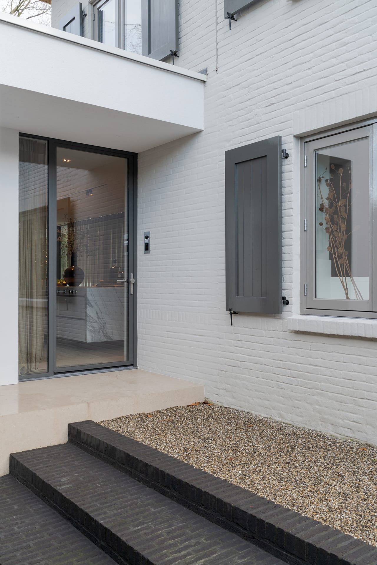 Entree van de villa met gevel in witte KEIM-verf, grijze luiken en een glazen deur met zicht op het marmeren keukeneiland.